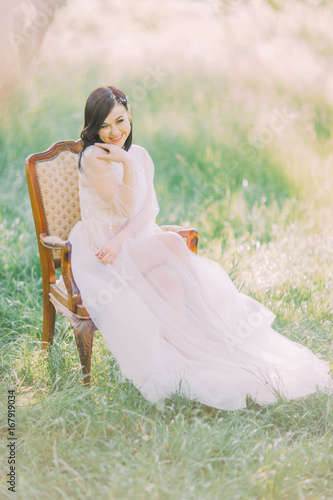 The full-lenght photo of the smiling woman in long white dress sitting on the old modern chair in the green spring field. photo