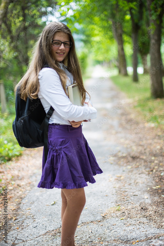 Pretty beautiful blonde child schoolgirl happily smiling and enjoying life coming back to school through alley in the nature with backpack, hugging book with hands, glasses, purple skirt and shirt Pretty beautiful blonde child schoolgirl happily smiling and enjoying life coming back to school through alley in the nature with backpack, hugging book with hands, glasses, purple skirt and shirt