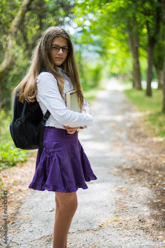 Pretty beautiful blonde child schoolgirl posing and enjoying life coming back to school through alley in the nature with backpack, hugging book with hands, glasses, purple skirt and shirt Pretty beautiful blonde child schoolgirl posing and enjoying life coming back to school through alley in the nature with backpack, hugging book with hands, glasses, purple skirt and shirt