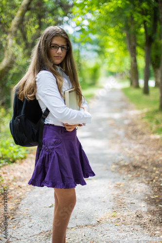 Pretty beautiful blonde child schoolgirl posing and enjoying life coming back to school through alley in the nature with backpack, hugging book with hands, glasses, purple skirt and shirt © HappyLenses