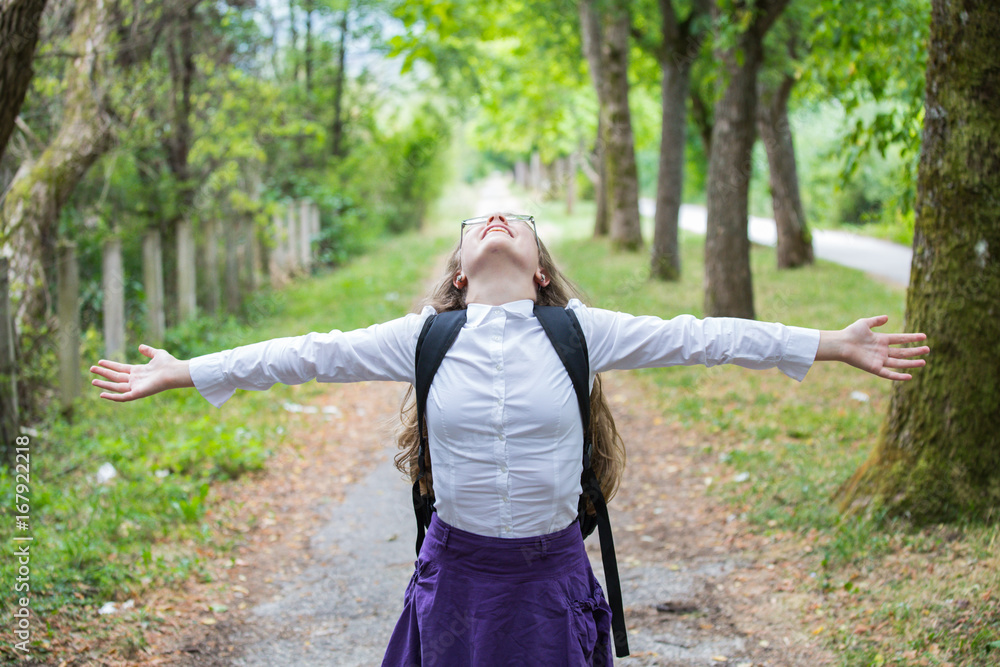 Beautiful pretty blonde school girl child with glasses, white shirt, purple skirt and backpack enjoying in the nature alley with arms widespread happily back to school Beautiful pretty blonde school girl child with glasses, white shirt, purple skirt and backpack enjoying in the nature alley with arms widespread happily back to school