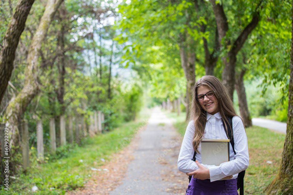 Pretty beautiful blonde child schoolgirl smiling holding notebooks enjoying back to school walking through alley in the nature with backpack, glasses, white shirt and purple skirt in the autumn Pretty beautiful blonde child schoolgirl smiling holding notebooks enjoying back to school walking through alley in the nature with backpack, glasses, white shirt and purple skirt in the autumn