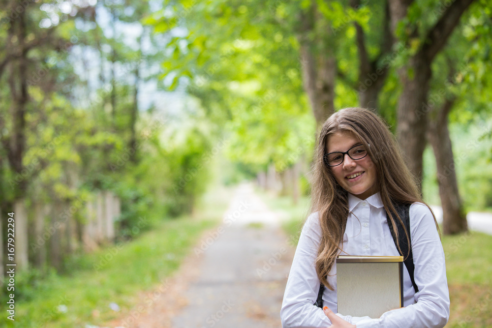 Pretty beautiful blonde child schoolgirl portrait smiling holding notebooks enjoying back to school walking through alley in the nature with backpack, glasses and white shirt in the autumn Pretty beautiful blonde child schoolgirl portrait smiling holding notebooks enjoying back to school walking through alley in the nature with backpack, glasses and white shirt in the autumn
