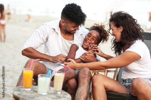 Young mixed race family sitting and relaxing  at the beach on beautiful summer day.Daughter lies in father lap and laughing.