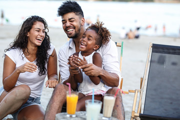 Young mixed race family sitting and relaxing  at the beach on beautiful summer day.Daughter lies in father lap and laughing.