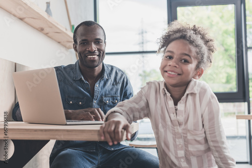 smiling african-american father and daughter sitting in cafe with laptop