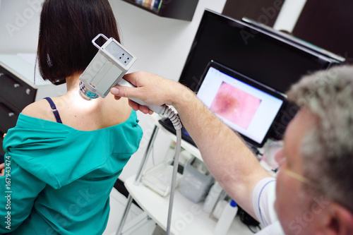 Canvas Print The doctor examines neoplasms or moles on the patient's skin - a young girl using a special apparatus for dermatoscopy - a dermoscope