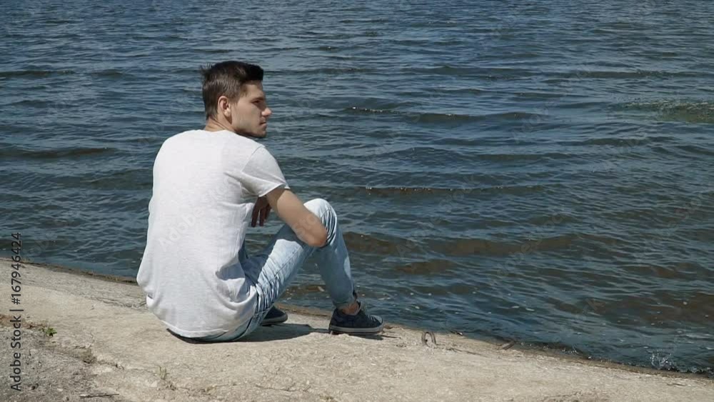 Young man sitting alone at bank of the river looking at water. People stock footage shot at summer season time. Young man sitting alone at bank of the river looking at water. People stock footage shot at summer season time.