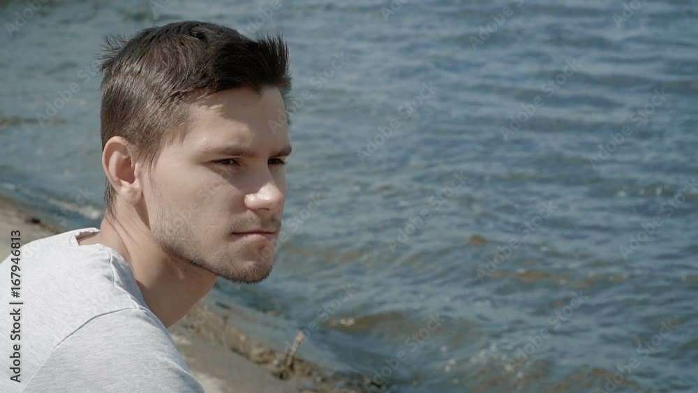 Young man sitting alone at bank of the river looking at water. People stock footage shot at summer season time. Young man sitting alone at bank of the river looking at water. People stock footage shot at summer season time.