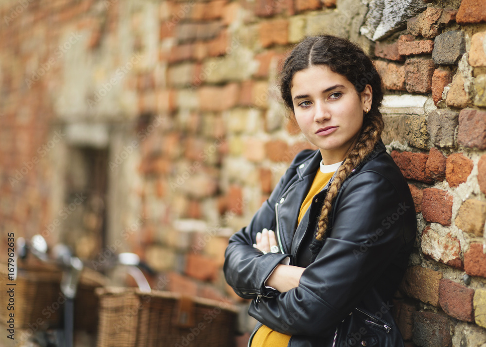 Young girl wearing motorbike jacket leans her back against ancient brick wall in italian street Young girl wearing motorbike jacket leans her back against ancient brick wall in italian street