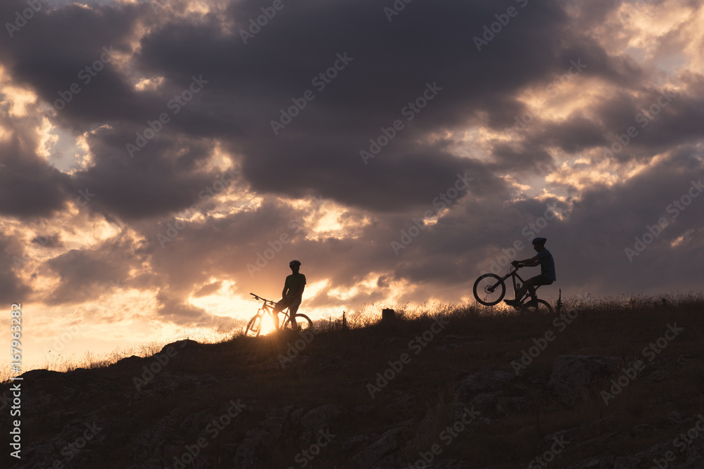 Two male bikers standing a hill admiring the sunset Two male bikers standing a hill admiring the sunset