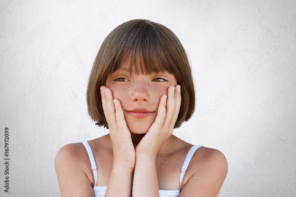 Close up portrait of adorable freckled girl with bobbed hairstyle, keeping her hands on cheeks, having dreamy expression, dreaming about new toy or holidays on sea. Little child with thoughtful look Close up portrait of adorable freckled girl with bobbed hairstyle, keeping her hands on cheeks, having dreamy expression, dreaming about new toy or holidays on sea. Little child with thoughtful look