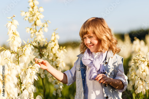 A portrait of a cute little boy with blue eyes and long blond hair in a jeans west outside at sunset in the field of white yucca flowers having fun with raised hands up