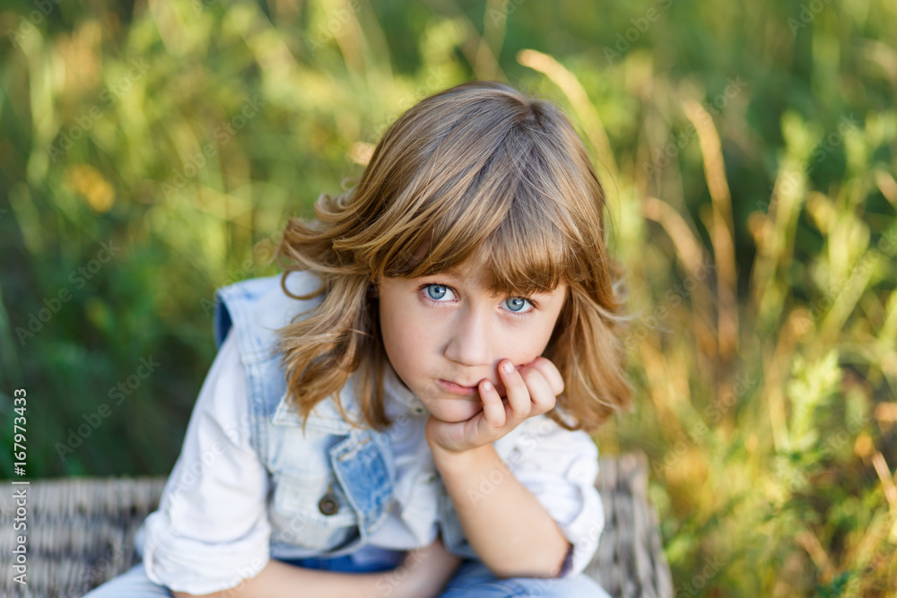 A portrait of a cute little boy with blue eyes and long blond hair in a jeans west sitting on a basket outside at sunset an having fun