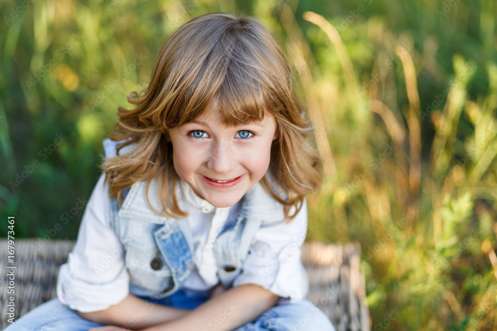 A portrait of a cute little boy with blue eyes and long blond hair in a jeans west sitting on a basket outside at sunset an having fun A portrait of a cute little boy with blue eyes and long blond hair in a jeans west sitting on a basket outside at sunset an having fun