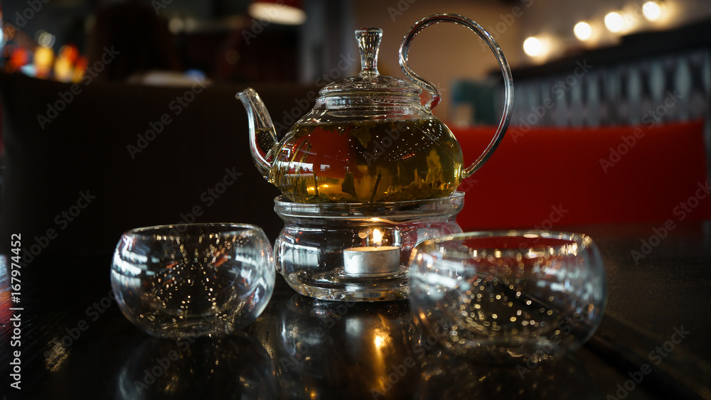 Two glass cups of herbal tee stay on a dark black wood table. Behind of caps a beautifull candle under a glass cover. Everything is in deep dark colors under a warm light. Romantic time. Two glass cups of herbal tee stay on a dark black wood table. Behind of caps a beautifull candle under a glass cover. Everything is in deep dark colors under a warm light. Romantic time.