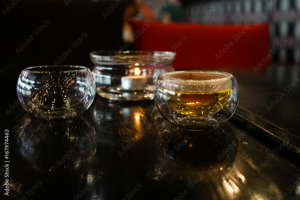 Two glass cups of herbal tee stay on a dark black wood table. Behind of caps a beautifull candle under a glass cover. Everything is in deep dark colors under a warm light. Romantic time. 