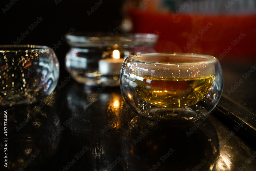 Two glass cups of tee stay on a dark black wood table. Behind of caps a beautifull candle under a glass cover. Everything is in deep dark colors under a warm light. Close up picture. Two glass cups of tee stay on a dark black wood table. Behind of caps a beautifull candle under a glass cover. Everything is in deep dark colors under a warm light. Close up picture.