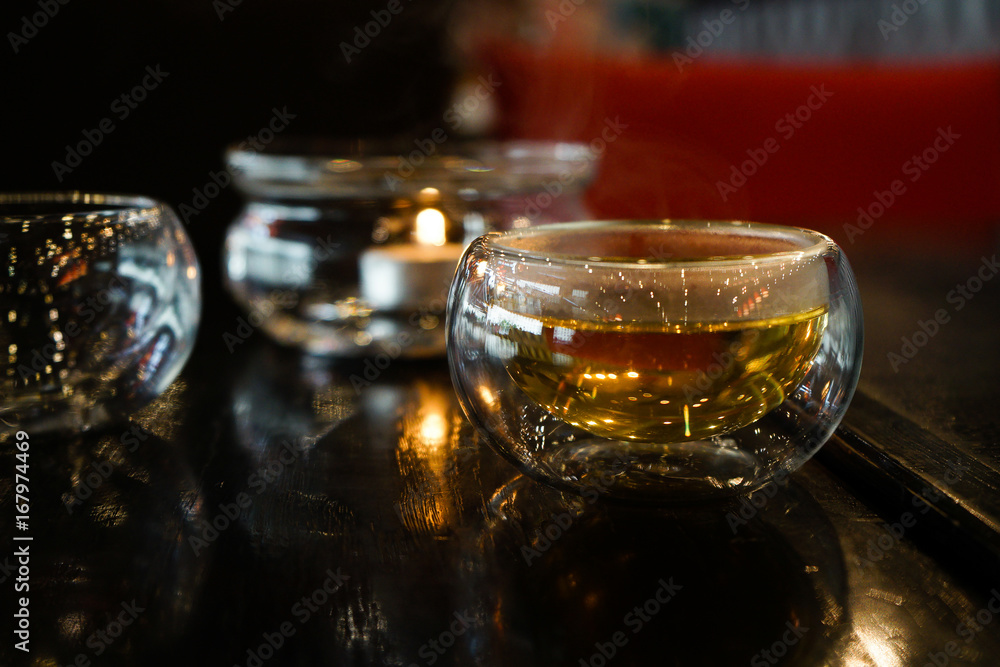 Two glass cups of tee stay on a dark black wood table. Behind of caps a beautifull candle under a glass cover. Everything is in deep dark colors under a warm light. Close up picture. Two glass cups of tee stay on a dark black wood table. Behind of caps a beautifull candle under a glass cover. Everything is in deep dark colors under a warm light. Close up picture.
