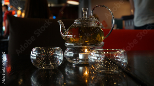 Two glass cups of herbal tee stay on a dark black wood table. Behind of caps a beautifull candle under a glass cover. Everything is in deep dark colors under a warm light. Romantic time.
