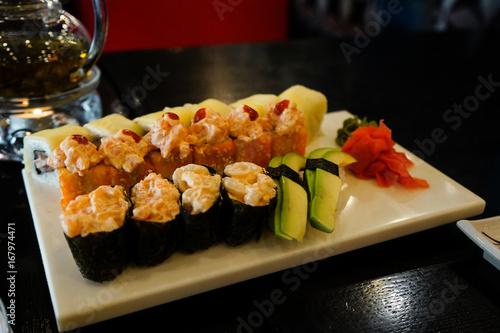 Different type of tasty sushi are puted on a big white square plate. It is all on a black table. In left corner is circle candle. Everything is in deep dark colors under a warm light.