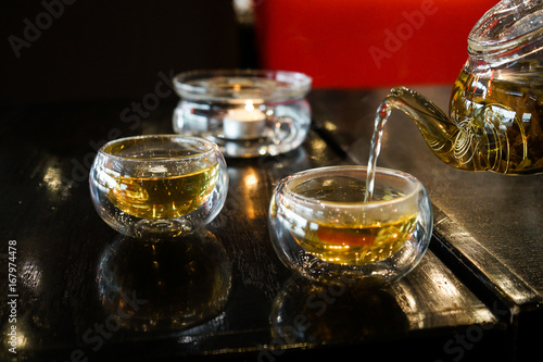 Two glass cups of herbal tee stay on a dark black wood table. Everything is in deep dark colors under a warm light. Romantic time. Water fills the cups from teapot.