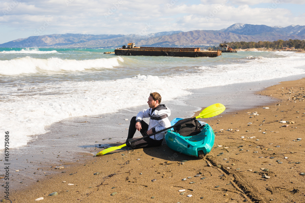 Young caucasian man sitting on beach with kayak Young caucasian man sitting on beach with kayak