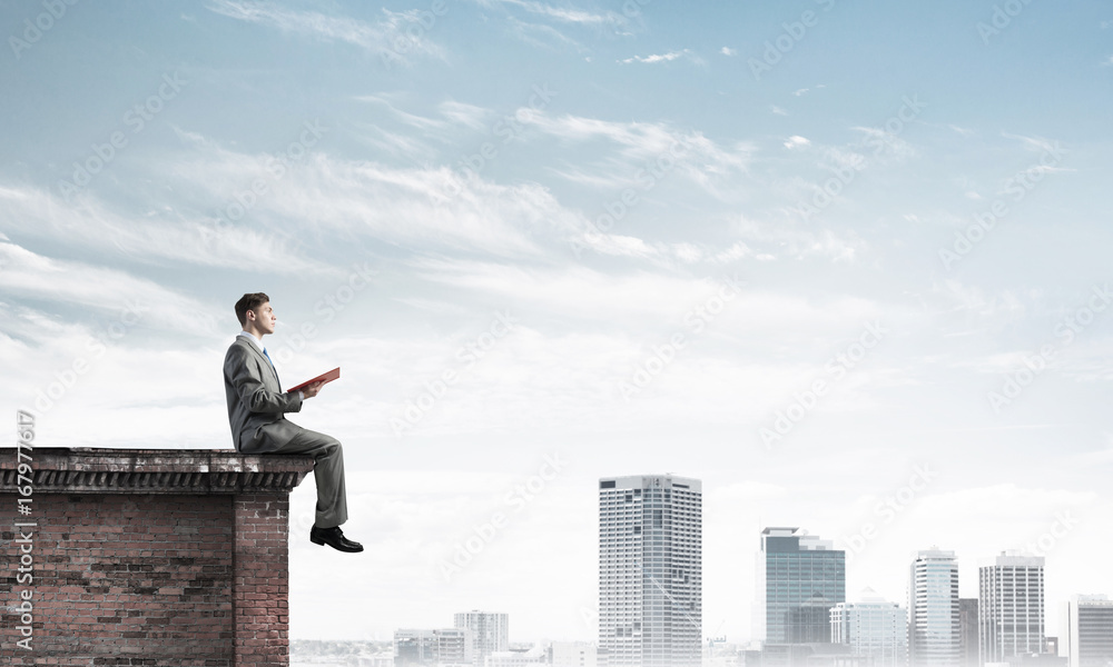 Man on roof edge reading book and cityscape at background Man on roof edge reading book and cityscape at background