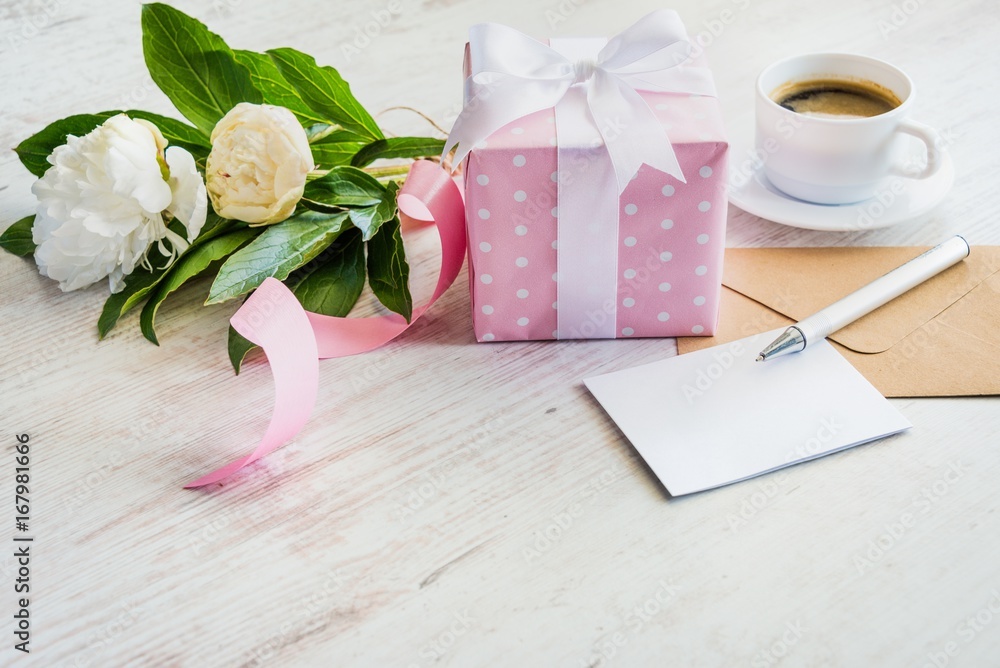 Pink dotted gift box, empty greeting card, kraft envelope, peonies bouquet and coffee cup over white wooden rustic table. Romantic holiday and event concept.