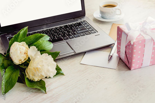 Side view of a deck with computer, bouquet of peonies flowers, cup of coffee, empty card and pink dotted gift box. White rustic wooden background. Romantic and feminine outlook.
