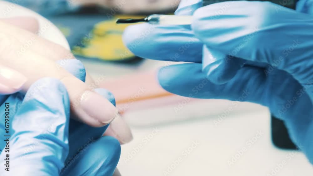 Cosmetician hand in blue rubber gloves puts transparent nail polish using brush on client thumb nail in bright salon, close up Cosmetician hand in blue rubber gloves puts transparent nail polish using brush on client thumb nail in bright salon, close up