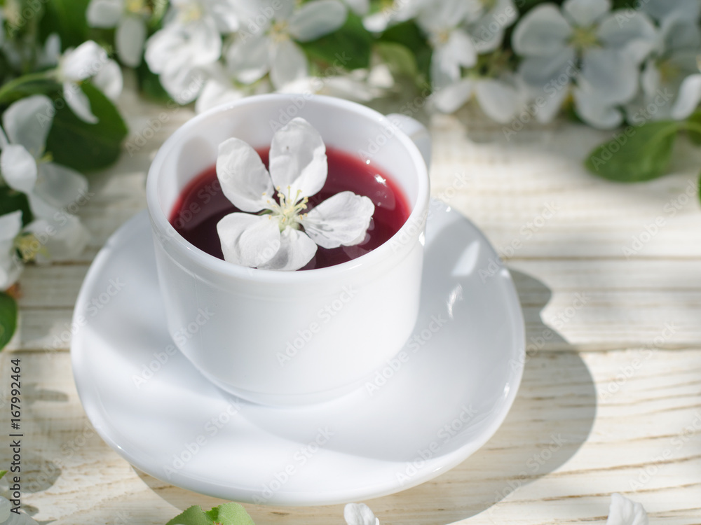 White mug of tea on a wooden table, apple blossoms in the background. Sunny, side view