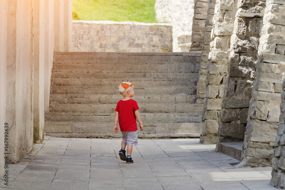 Little boy walking alone among the ancient walls, sunlight. Back view Little boy walking alone among the ancient walls, sunlight. Back view