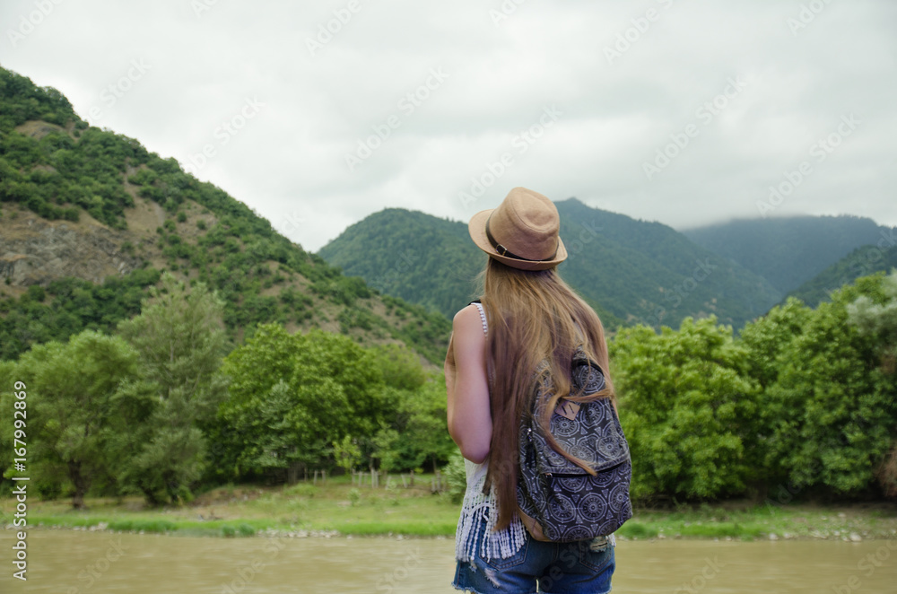 Girl with a backpack and a hat admiring the green mountains and the river. View from the back