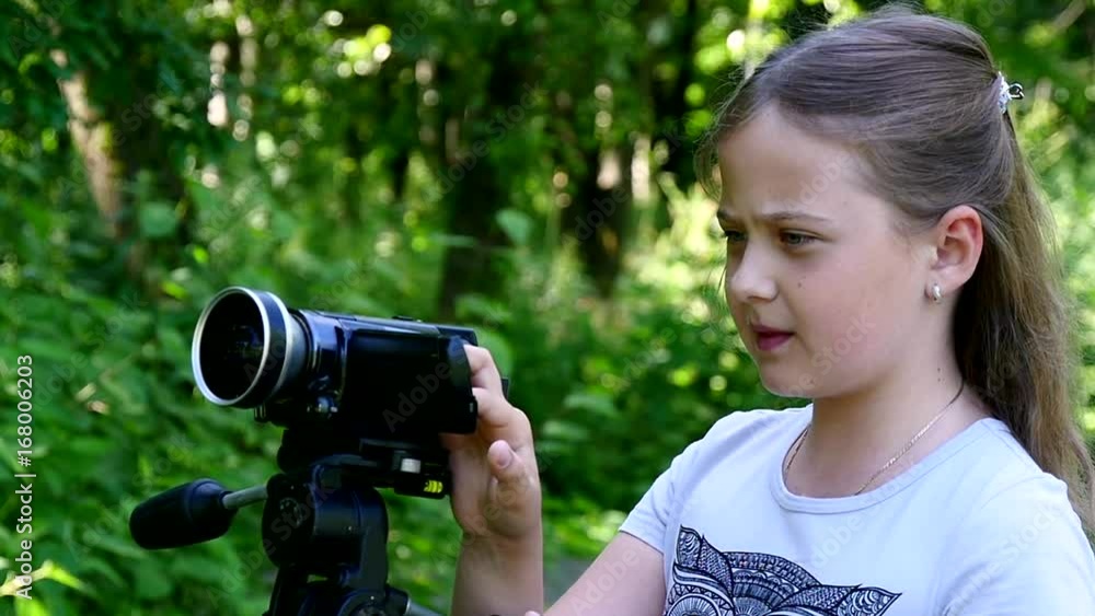 Young girl with video camera shoots movies in nature on green park slow motion. Kids outdoors in summer are creative work of cinema. Interesting to look at world in childhood. Young girl with video camera shoots movies in nature on green park slow motion. Kids outdoors in summer are creative work of cinema. Interesting to look at world in childhood.