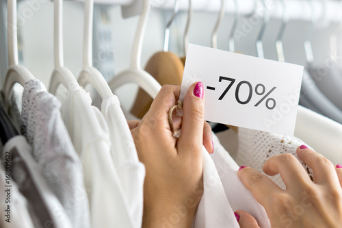 Female customer browsing clothes in a shop. Woman shopping for fashion offer and deal. Holding price tag with minus seventy percent or 70% sale, bargain and reduced cheap prices in clothing store. photo