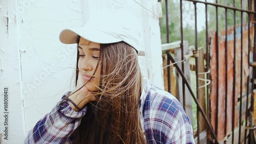 A young woman with long hair and a baseball cap is standing by the old fence.