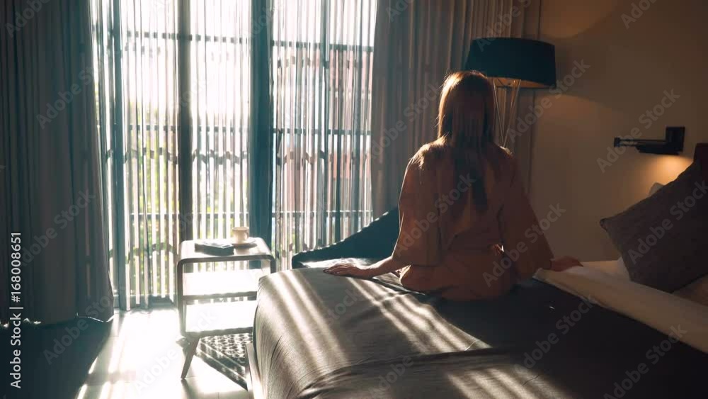 Young caucasian woman in brown beige robe sitting on neat bed in the morning in front of big window. Shot with Sony a7s and Atomos Ninja Flame.