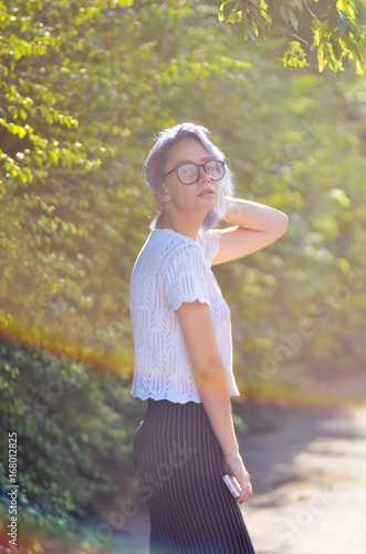 Fotografija Young informal hipster girl with ashen purple hair in glasses posing in front of a camera in the park on a background of green bushes