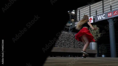 Blonde with long hair in a red skirt and a black topic makes twine in the dance in the middle of a shopping center, the view from under the bench