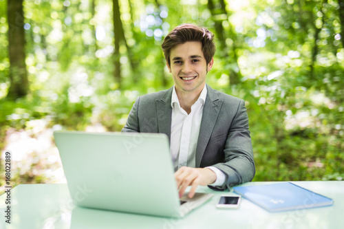 Businessman working on laptop computer in the forest at office table and smile at camera. Business concept.