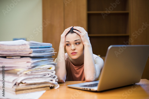 Fotografie A young woman sits in front of a pile of papers and a computer holding her head