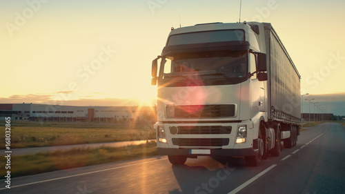 Front-View of Semi-Truck with Cargo Trailer Driving on a Highway. He s Speeding Through Industrial Warehouse Area with Sunset in the Background.