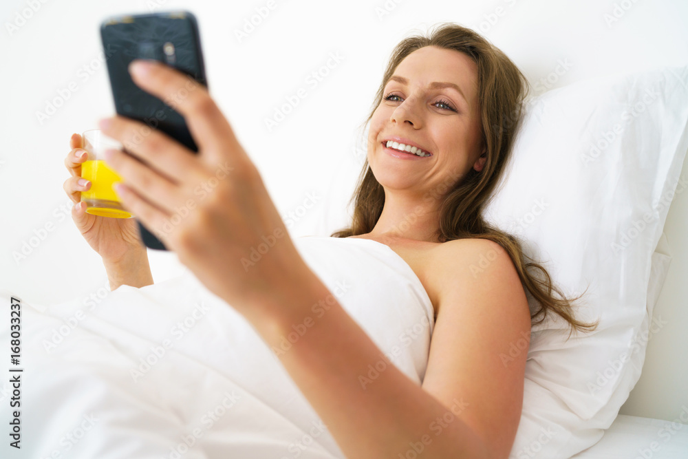Cute woman checks the smartphone and drinks orange juice in bed in the morning Cute woman checks the smartphone and drinks orange juice in bed in the morning