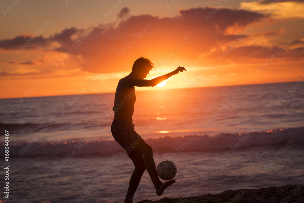 Young male playing football on the beach at sunset