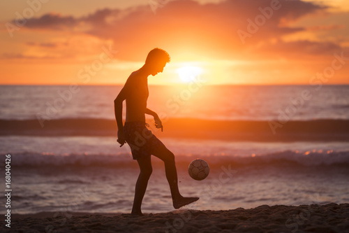 Young male playing football on the beach at sunset