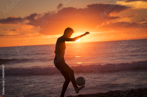 Young male playing football on the beach at sunset