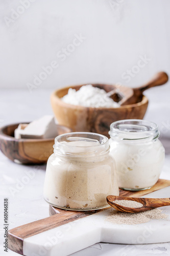 Rye and wheat sourdough in glass jars, fresh and instant yeast, olive wood bowl of flour for baking homemade bread. With spoon, serving board over gray concrete background.