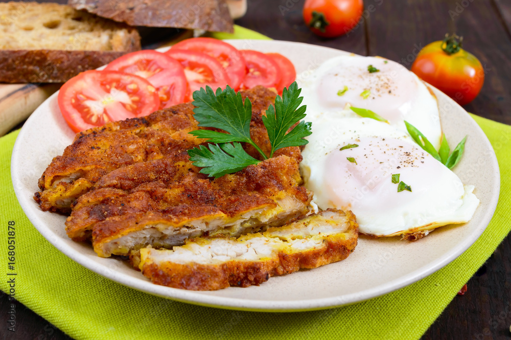 Pieces of chop (schnitzel), toast with eggs, fresh tomato on a dark wooden background.