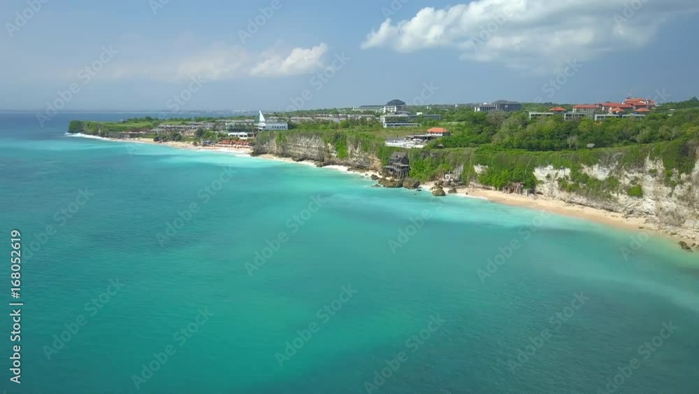 AERIAL: Flying along oceanfront villas, bungalows, holiday houses & cabins in luxury resort on cliffy coastline on lush green paradise island. People swimming in sea, waves washing sandy beach in Bali